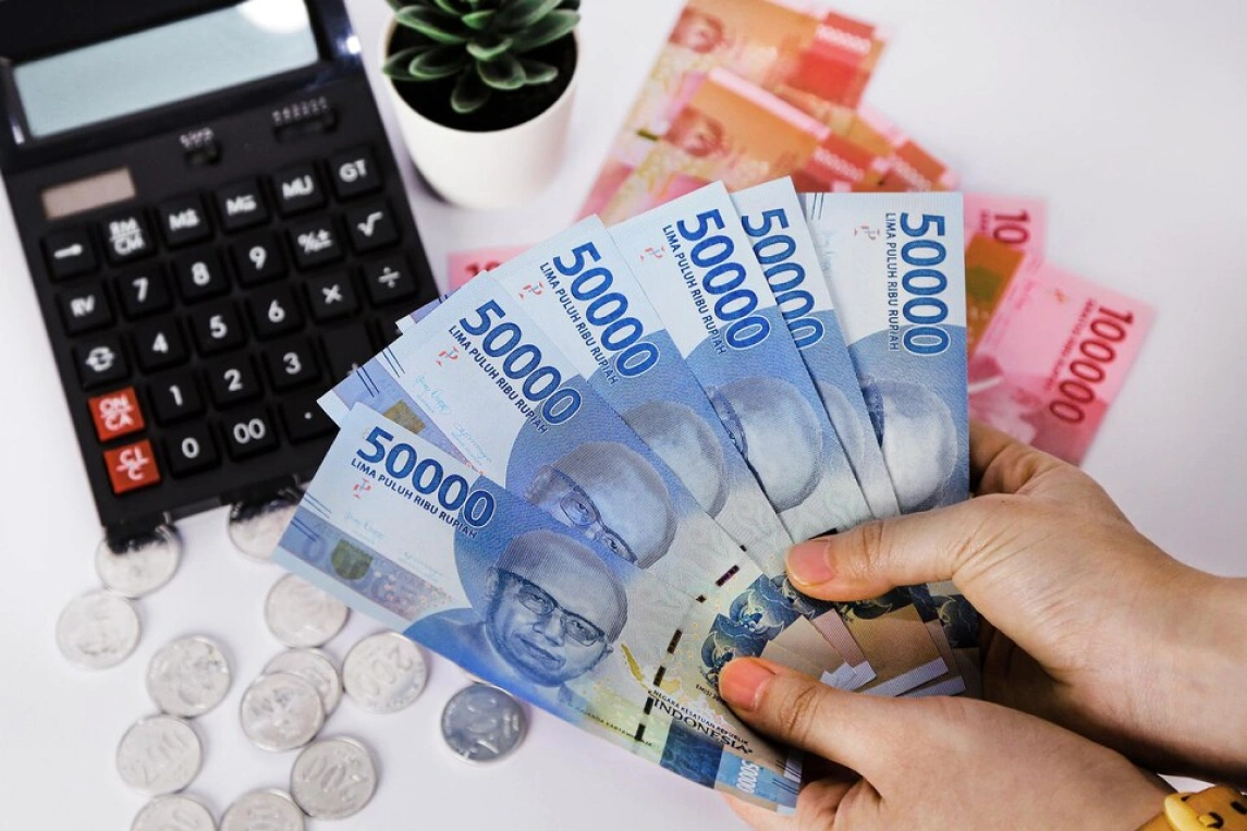 Tangan Memegang Uang Rupiah Dengan Kalkulator Dan Uang Koin Di Latar Belakang.